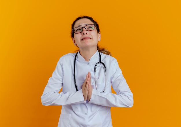 Foto gratuita mirando hacia arriba preocupada joven doctora vistiendo bata médica y estetoscopio con gafas mostrando gesto de oración aislado