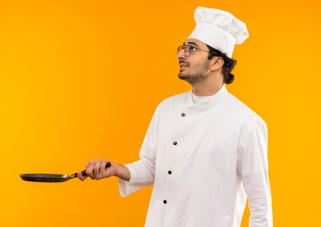 Mirando hacia arriba preocupado joven cocinero vistiendo uniforme de chef y gafas sosteniendo una sartén