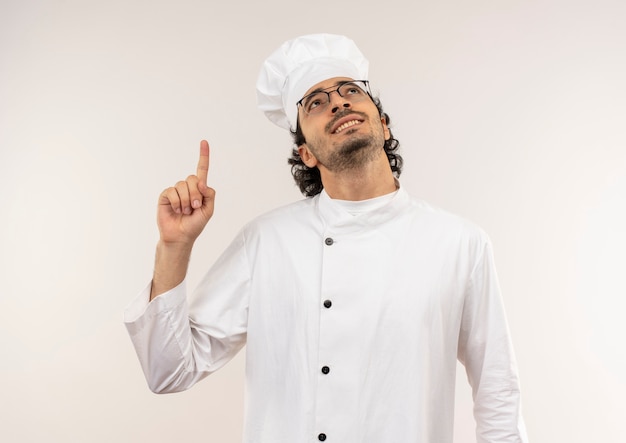 Mirando hacia arriba sonriente joven cocinero vistiendo uniforme de chef y gafas apunta a un lado