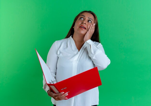 Mirando hacia arriba sorprendida mujer de mediana edad caucásica casual sosteniendo la carpeta y poniendo la mano en la mejilla