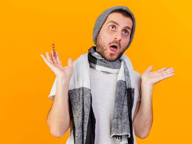 Mirando hacia arriba sorprendido joven enfermo vestido con gorro y bufanda de invierno sosteniendo pastillas y extendiendo las manos aisladas sobre fondo amarillo