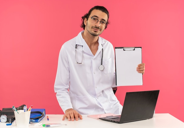 Mirando a la cámara complacido joven médico con gafas médicas vistiendo bata médica con estetoscopio de pie detrás del escritorio