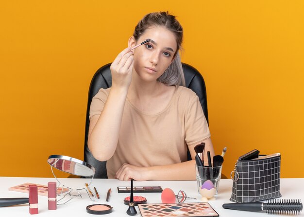 Mirando a la cámara hermosa joven se sienta a la mesa con herramientas de maquillaje aplicando sombra de ojos con pincel de maquillaje aislado en la pared naranja