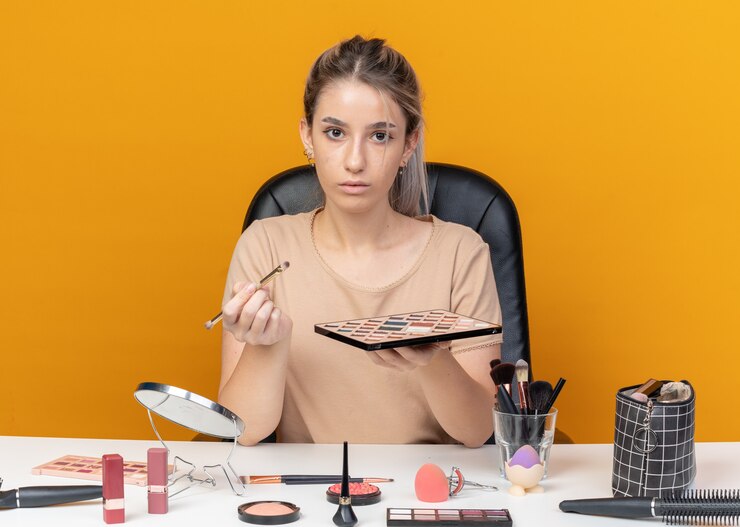 Foto gratuito mirando a la cámara hermosa joven se sienta a la mesa con herramientas de maquillaje sosteniendo prush con paleta de sombra de ojos aislada en la pared naranja