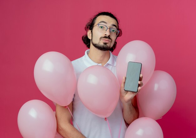 Mirando a la cámara hombre guapo con gafas sosteniendo globos y teléfono aislado sobre fondo rosa