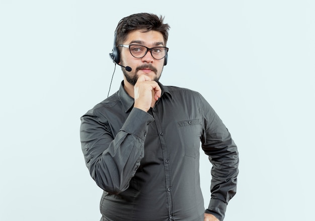 Mirando a la cámara joven centro de llamadas hombre con auriculares y gafas poniendo la mano en el mentón aislado sobre fondo blanco.