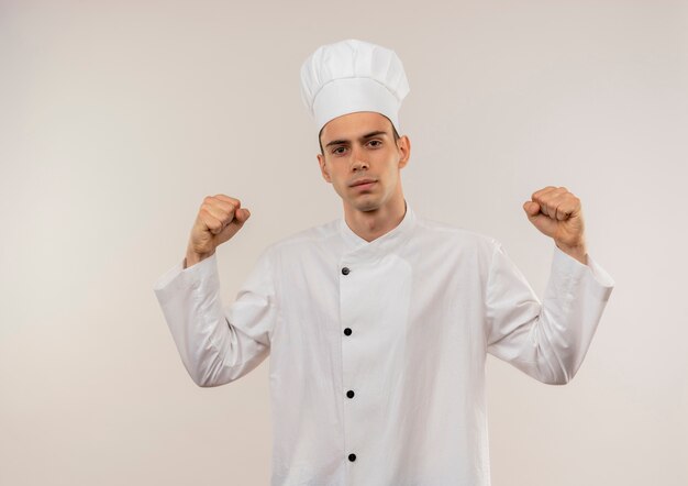 Mirando a la cámara joven cocinero con uniforme de chef haciendo un gesto fuerte en la pared blanca aislada con espacio de copia