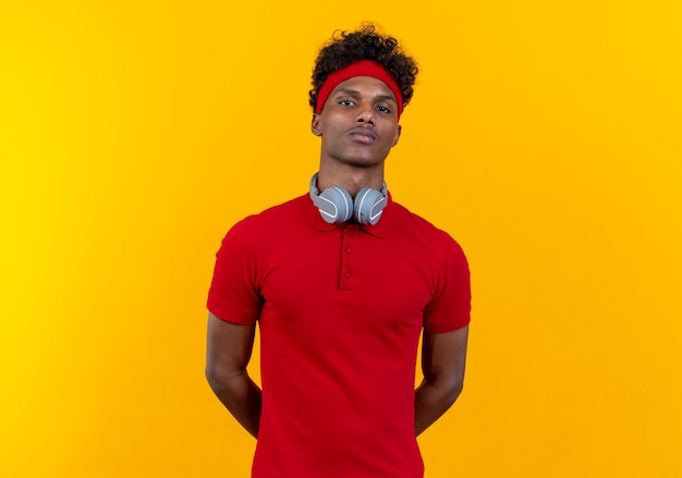 Mirando a la cámara joven deportivo afroamericano con diadema y muñequera con auriculares manteniendo las manos detrás de la espalda aislado sobre fondo amarillo