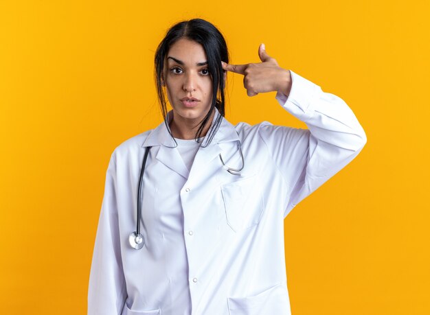 Mirando a la cámara joven doctora vistiendo bata médica con estetoscopio poniendo el dedo en la sien aislado en la pared amarilla