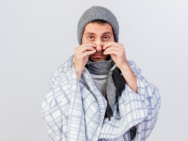 Mirando a la cámara joven enfermo con gorro de invierno y bufanda envuelto en plaid poniendo yeso en la nariz aislado en blanco