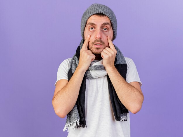 Mirando a la cámara joven enfermo con sombrero de invierno con bufanda abriendo los ojos con los dedos aislados sobre fondo púrpura