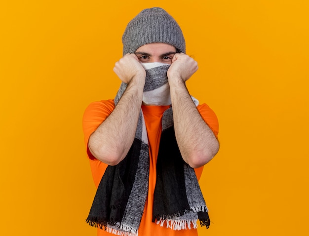 Foto gratuita mirando a la cámara joven enfermo vistiendo gorro de invierno con bufanda cubrió la cara con bufanda aislado sobre fondo naranja