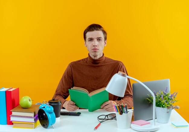 mirando a la cámara joven estudiante chico sentado en el escritorio con herramientas escolares sosteniendo el libro