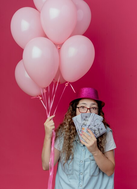 Mirando a la cámara joven con gafas y sombrero rosa sosteniendo globos y boca cubierta con dinero en efectivo aislado sobre fondo rosa