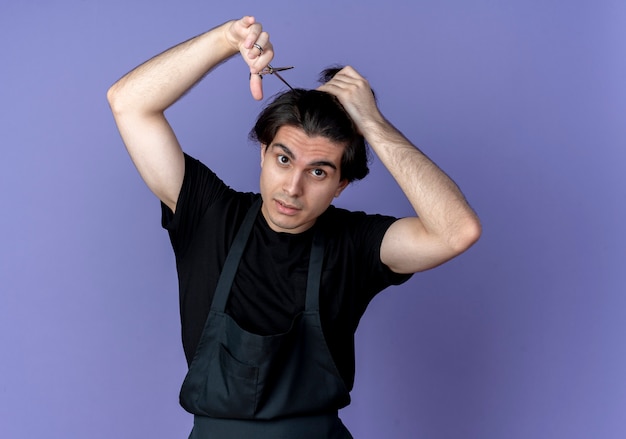 Mirando a la cámara joven guapo barbero en uniforme cortando el cabello con tijeras aislado sobre fondo azul.