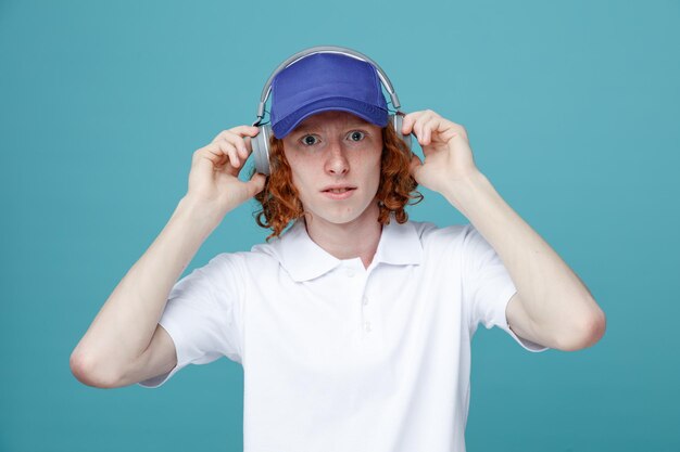 Mirando a la cámara joven guapo con gorra usando auriculares aislados en fondo azul