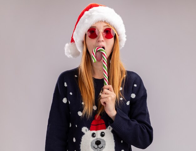 Mirando a la cámara joven hermosa niña con sombrero de navidad y gafas sosteniendo y lamiendo dulces navideños aislado sobre fondo blanco.
