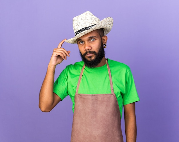 Mirando a la cámara joven jardinero chico afroamericano con sombrero de jardinería aislado en la pared azul