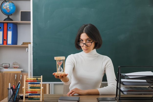 mirando a la cámara una joven maestra con gafas sosteniendo un reloj de arena sentada en un escritorio con herramientas escolares en el aula