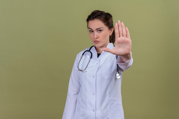 Mirando a la cámara joven médico con bata médica con estetoscopio muestra gesto de parada en la pared verde