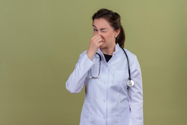 Foto gratuita mirando a la cámara joven médico vistiendo bata médica con estetoscopio nariz cerrada en la pared verde