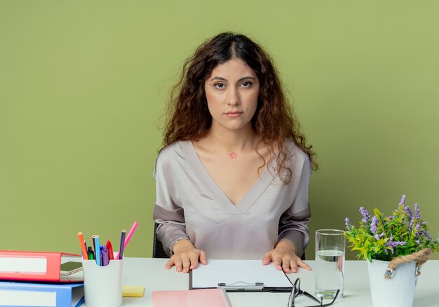 Mirando a la cámara joven oficinista bastante femenina sentada en un escritorio con herramientas de oficina aisladas sobre fondo verde oliva