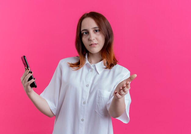 Mirando a la cámara joven pelirroja sosteniendo el teléfono y extendiendo la mano a la cámara aislada sobre fondo rosa