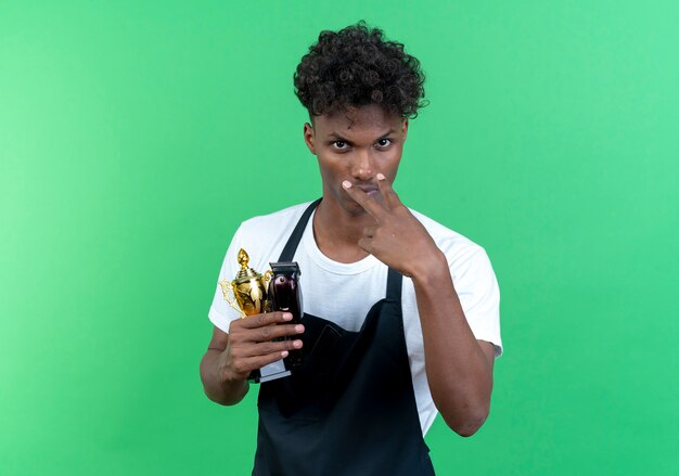 Mirando a la cámara joven peluquero afroamericano vistiendo uniforme sosteniendo la copa ganadora y mostrando mirándote gesto aislado sobre fondo verde