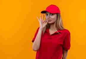 Foto gratuita mirando a la cámara joven repartidora vestida con uniforme rojo y gorra mostrando gesto okey aislado sobre fondo naranja
