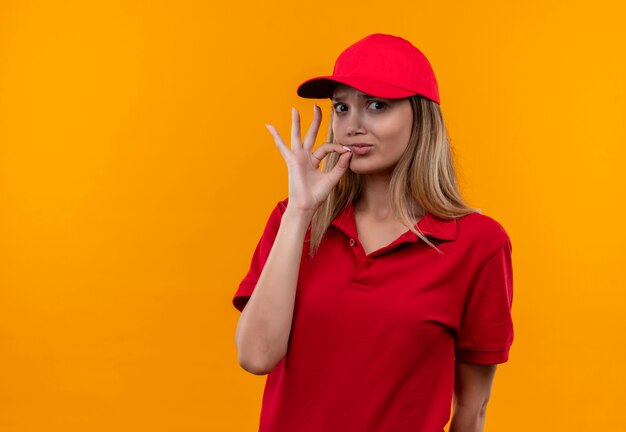 Mirando a la cámara joven repartidora vestida con uniforme rojo y gorra mostrando gesto okey aislado sobre fondo naranja