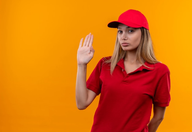 Mirando a la cámara joven repartidora vestida con uniforme rojo y gorra mostrando gesto de parada aislado sobre fondo naranja