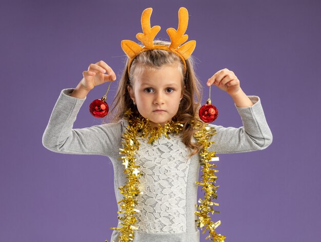 Mirando a la cámara niña vistiendo un aro de pelo de navidad con guirnalda en el cuello sosteniendo bolas de navidad aislado sobre fondo azul.