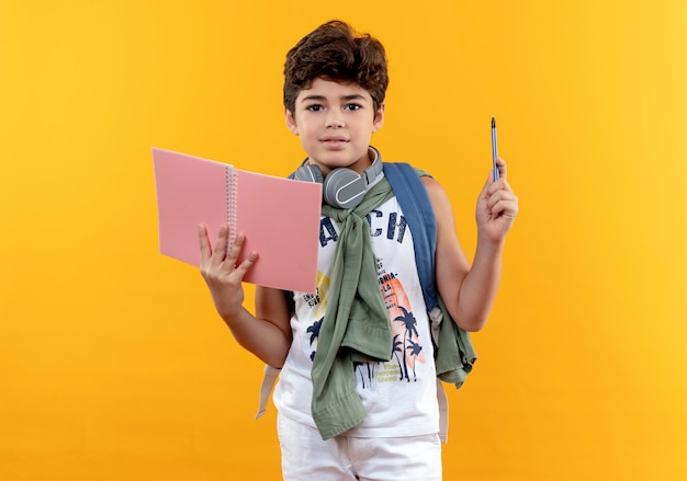 Mirando a la cámara niño de la escuela con mochila y auriculares sosteniendo un cuaderno y un bolígrafo aislado sobre fondo amarillo