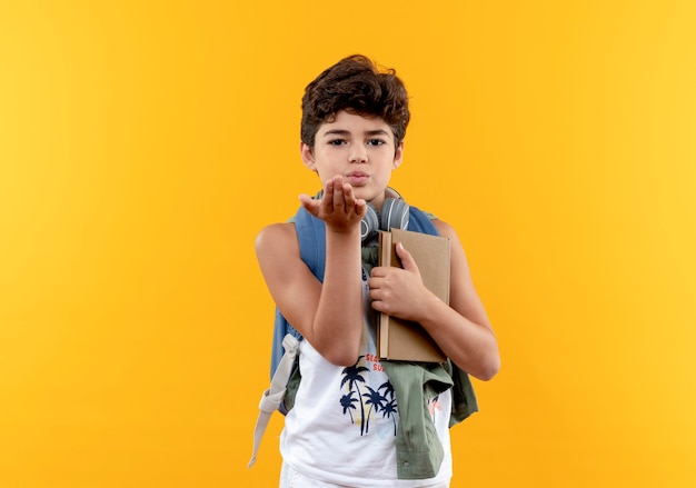 Mirando a la cámara niño de escuela con mochila y auriculares sosteniendo el libro y mostrando gesto de beso aislado sobre fondo amarillo