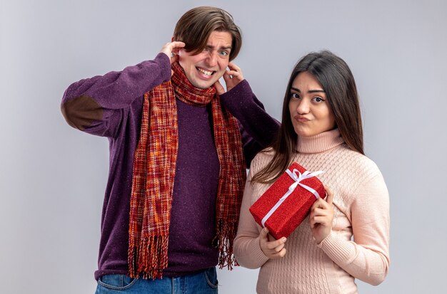 Mirando a la cámara pareja joven en el día de San Valentín chico oídos cerrados chica descontenta sosteniendo caja de regalo aislado sobre fondo blanco.