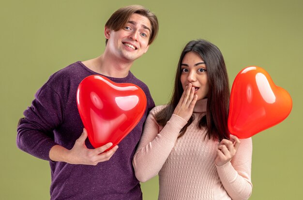 Mirando a la cámara pareja joven en el día de San Valentín sosteniendo globos de corazón aislado sobre fondo verde oliva