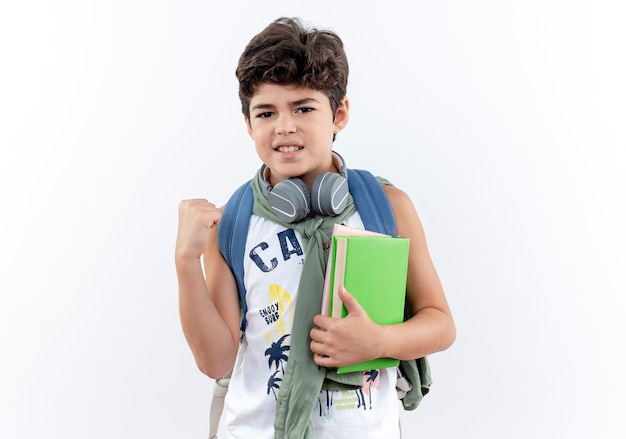 Mirando a la cámara pequeño colegial con mochila y auriculares sosteniendo el libro y cruzando el dedo aislado sobre fondo blanco.