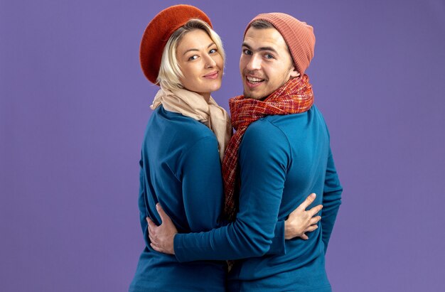 Mirando a la cámara sonriendo de pie detrás de la vista de la joven pareja con sombrero con bufanda en el día de San Valentín aislado sobre fondo azul.