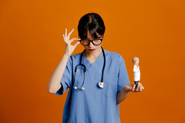 Mirando a la cámara sosteniendo el juguete joven doctora vistiendo uniforme fith estetoscopio aislado sobre fondo naranja