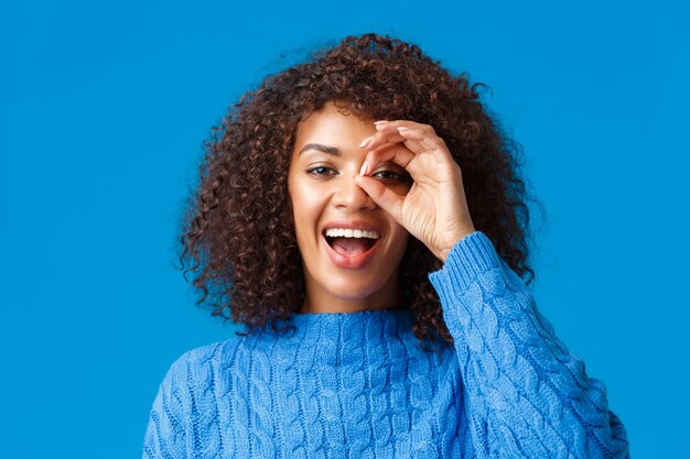 Mirando a la distancia. Close-up alegre atractiva mujer afroamericana en busca de algo, encontró un gran descuento de vacaciones, mire a través del letrero bien y sonriendo complacido, pared azul.