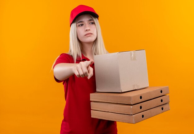 Mirando a la entrega lateral joven vistiendo camiseta roja y gorra sosteniendo la caja de pizza apunta al lado de la pared naranja aislada
