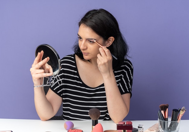 Mirando el espejo complacido mirando a la hermosa joven se sienta a la mesa con herramientas de maquillaje aplicando sombra de ojos con pincel de maquillaje aislado en la pared azul