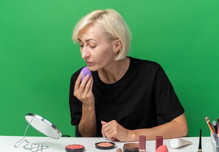 Foto gratuito mirando el espejo hermosa joven se sienta a la mesa con herramientas de maquillaje aplicando crema tonificante con esponja aislada en la pared verde