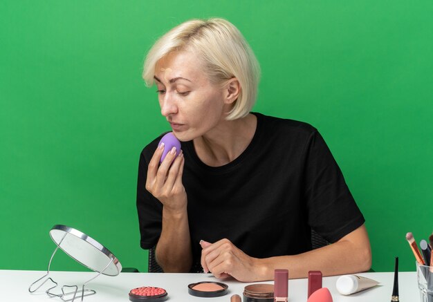 Mirando el espejo hermosa joven se sienta a la mesa con herramientas de maquillaje aplicando crema tonificante con esponja aislada en la pared verde