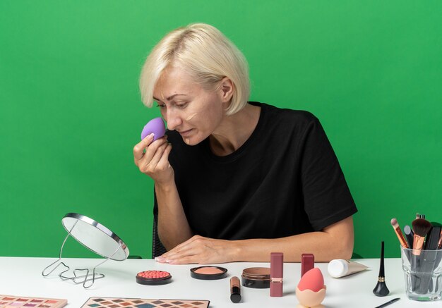 Mirando el espejo hermosa joven se sienta a la mesa con herramientas de maquillaje aplicando crema tonificante con esponja aislada en la pared verde