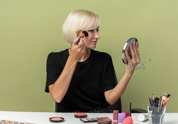 Foto gratuita mirando el espejo hermosa joven se sienta a la mesa con herramientas de maquillaje aplicando rubor en polvo aislado en la pared verde oliva