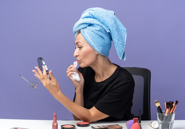 Mirando el espejo hermosa joven se sienta a la mesa con herramientas de maquillaje limpiando el cabello con una toalla limpiando la cara con una servilleta aislada en la pared azul