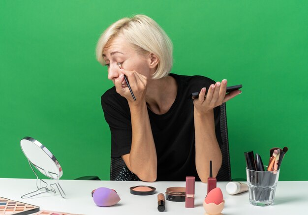 Mirando el espejo joven hermosa se sienta a la mesa con herramientas de maquillaje aplicando sombra de ojos con pincel de maquillaje aislado en la pared verde