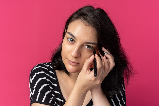 Mirando a la hermosa joven dibujar la flecha con delineador de ojos aislado en la pared rosa