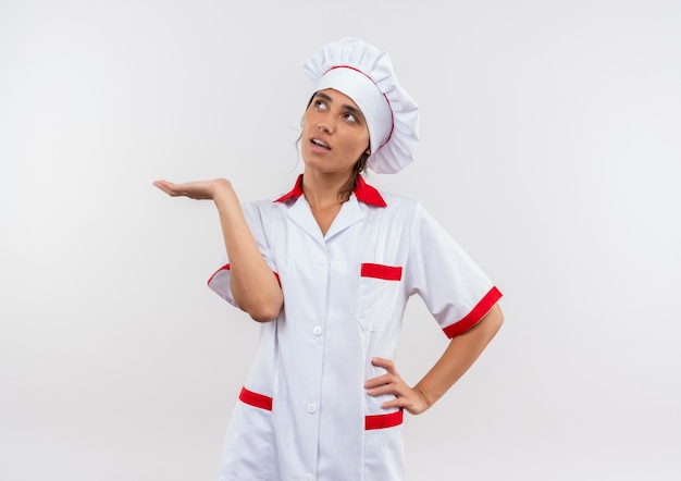 Mirando a la joven cocinera vistiendo uniforme de chef fingiendo sostener algo poniendo la mano en la cadera en la pared blanca aislada con espacio de copia